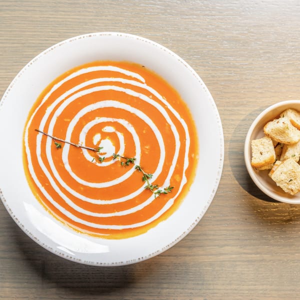 Overhead view of a bowl of orange soup with a white cream swirl and thyme, next to croutons.