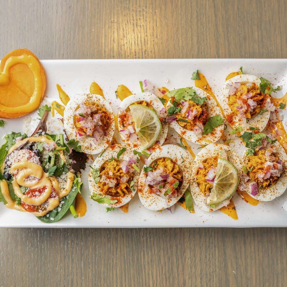 A white platter of Indian deviled eggs, garnished with spices, red onion, cilantro, and lime.
