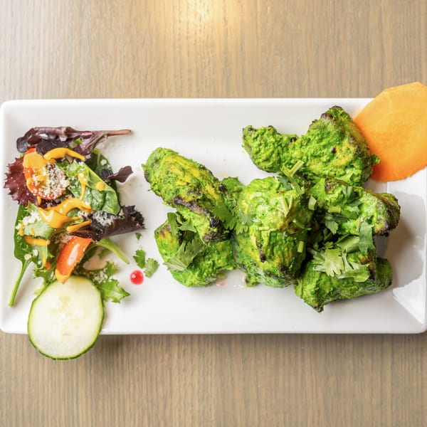 Green chicken tikka pieces with cilantro, a side salad, and a cucumber slice on a white plate.