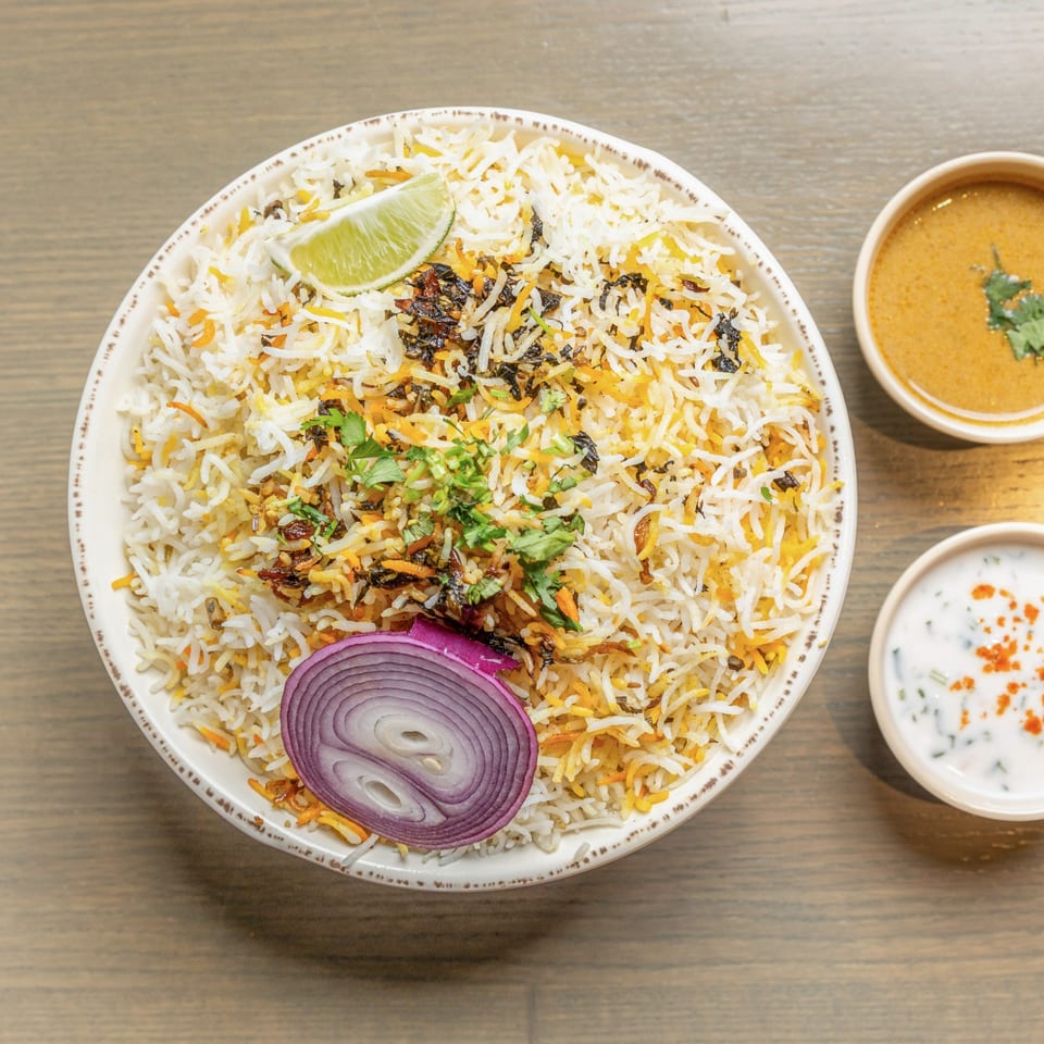 A large bowl of biryani rice with lime, red onion, and cilantro, served with two side sauces.